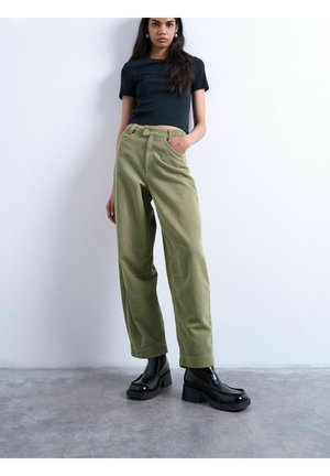 Ung kvinde iført sort kort crop-top, olivengrønne vidde corduroy bukser og sorte plateau ankelstøvler, stående mod en hvid væg.