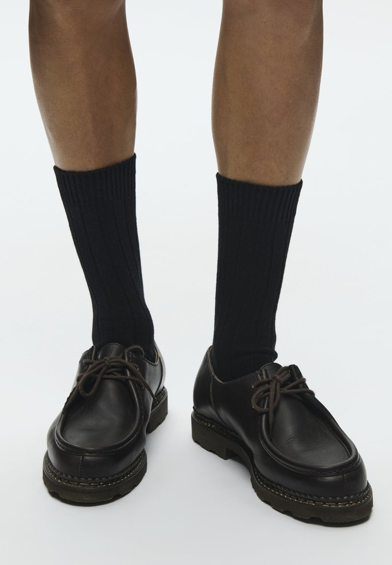 Jambes portant des chaussettes noires côtelées mi-mollet et des chaussures à lacets en cuir noir avec des semelles épaisses sur un fond clair.