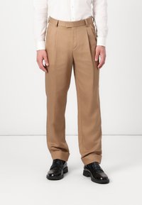 Beige tailored trousers with a straight fit, pleated front, cuffed hems, and belt loops, paired with black leather shoes.