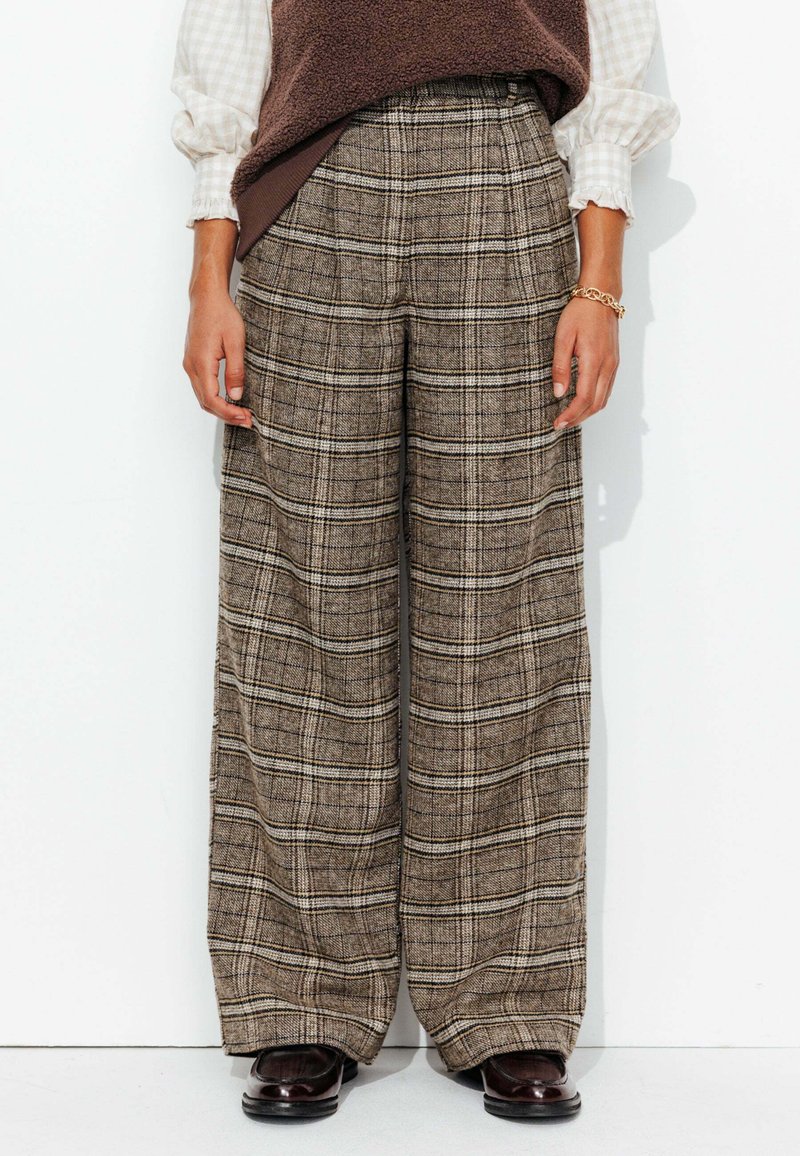 Pantalons à jambes larges en tissu écossais brun, présentant une surface texturée et un design taille haute. Complétés par des chaussures foncées.