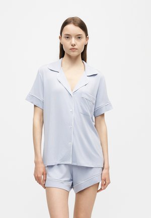 Jeune femme portant un haut de pyjama bleu clair à manches courtes avec col et boutonnières, et un short assorti, debout devant un fond uni.