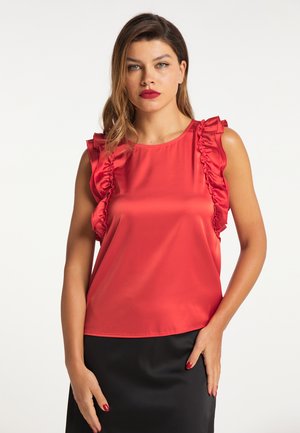 Vrouw met lang haar draagt een felrood mouwloos topje met ruches en een zwarte rok, staand tegen een effen lichte achtergrond.