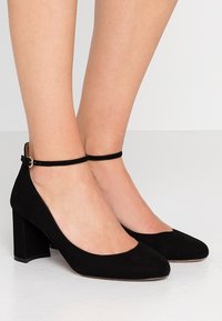 Schwarze Wildleder-Pumps mit hohen Absätzen, spitz zulaufender Zehenpartie, Knöchelriemen und Blockabsatz. Die Textur wirkt weich und hat ein mattes Finish.