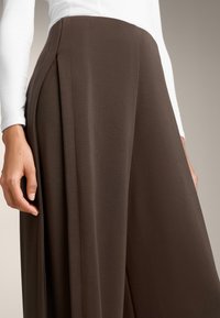 Pantaloni larghi marroni con texture, con pieghe e dettagli sulla cucitura laterale, abbinati a una maglietta bianca a maniche lunghe. Concentrati sul tessuto e sul taglio.