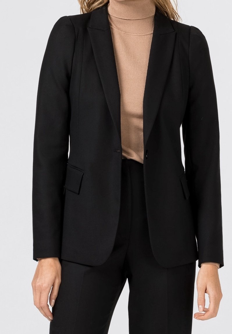 Femme portant un blazer noir taillé sur mesure et un pantalon noir par-dessus un pull à col roulé beige, debout devant un fond clair uni.
