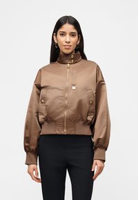 Γυναίκα που φοράει καφέ σατινέ bomber μπουφάν με χρυσά κουμπιά και φερμουάρ, συνδυασμένο με μαύρο παντελόνι σε λευκό φόντο.