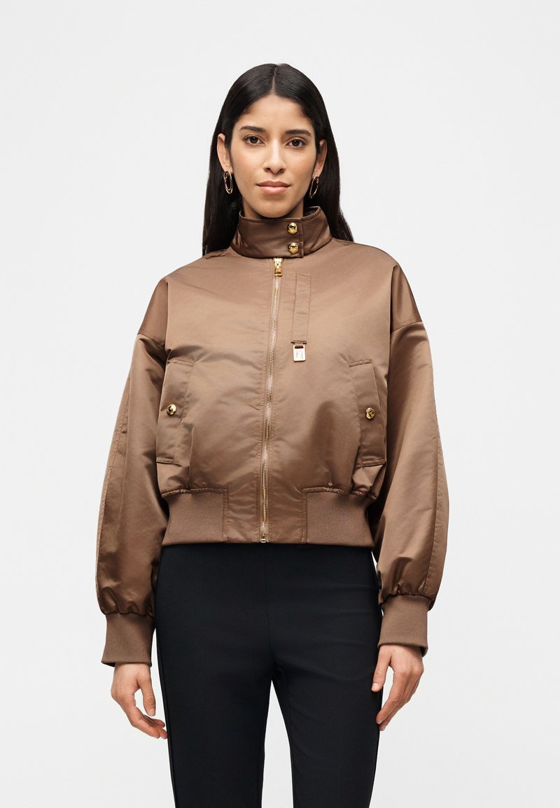 Γυναίκα που φοράει καφέ σατινέ bomber μπουφάν με χρυσά κουμπιά και φερμουάρ, συνδυασμένο με μαύρο παντελόνι σε λευκό φόντο.