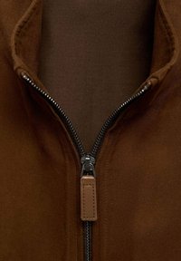 Chaqueta marrón con cremallera, hecha de un tejido grueso y texturizado, con una cremallera negra y un tirador beige, además de un forro interior suave.