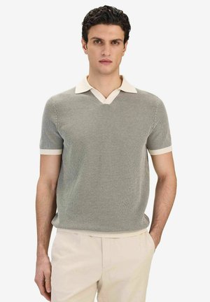 Man med mörkt hår som bär en grå kortärmad stickad pikétröja med beige krage och beige byxor, står mot en vit bakgrund.