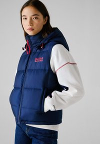 Μπλε γιλέκο με κουκούλα, με κόκκινες λεπτομέρειες, επώνυμο "RedBull Racing," και κλείσιμο με φερμουάρ. Φοριέται πάνω από μια λευκή φούτερ.