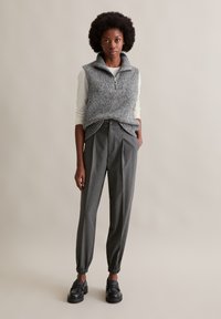 Frau steht und trägt eine graue gestrickte ärmellose Weste über einem weißen Langarmshirt, graue Nadelstreifen-Hose mit schmal zulaufendem Bein und schwarze Loafers.