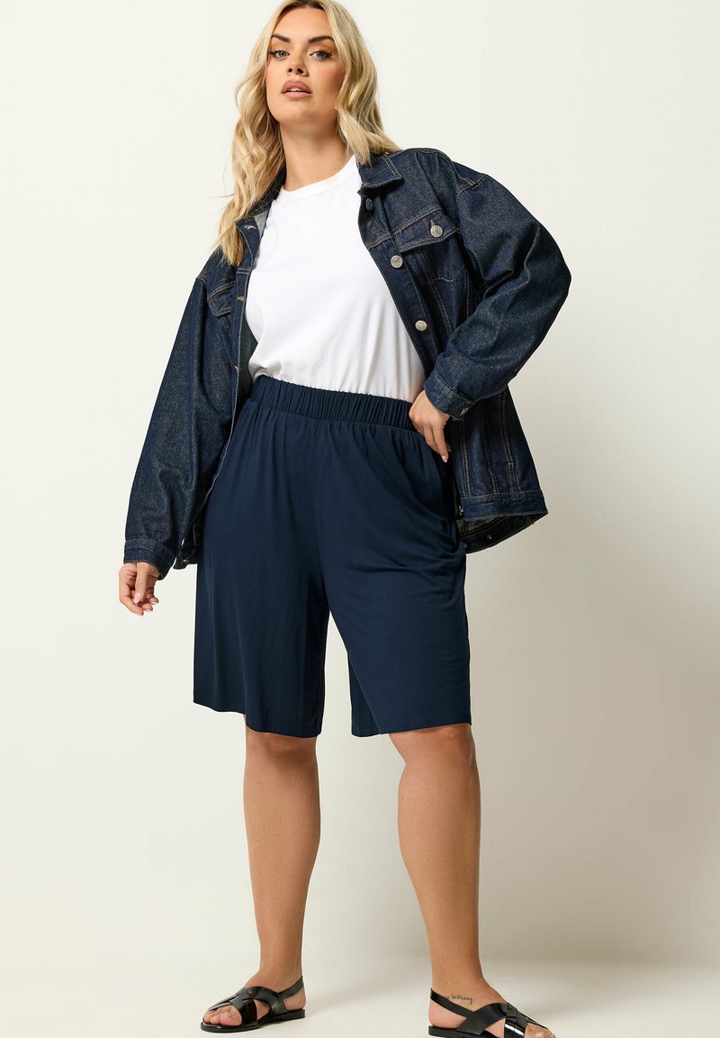 Femme blonde portant un t-shirt blanc, une veste en denim foncé, un short bleu marine à taille élastique et des sandales noires, posant devant un fond clair uni.