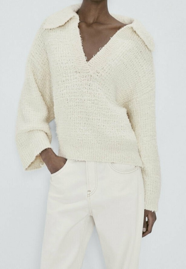 Personne portant un pull en maille texturée crème avec un col large et un pantalon blanc taille haute, main dans la poche.