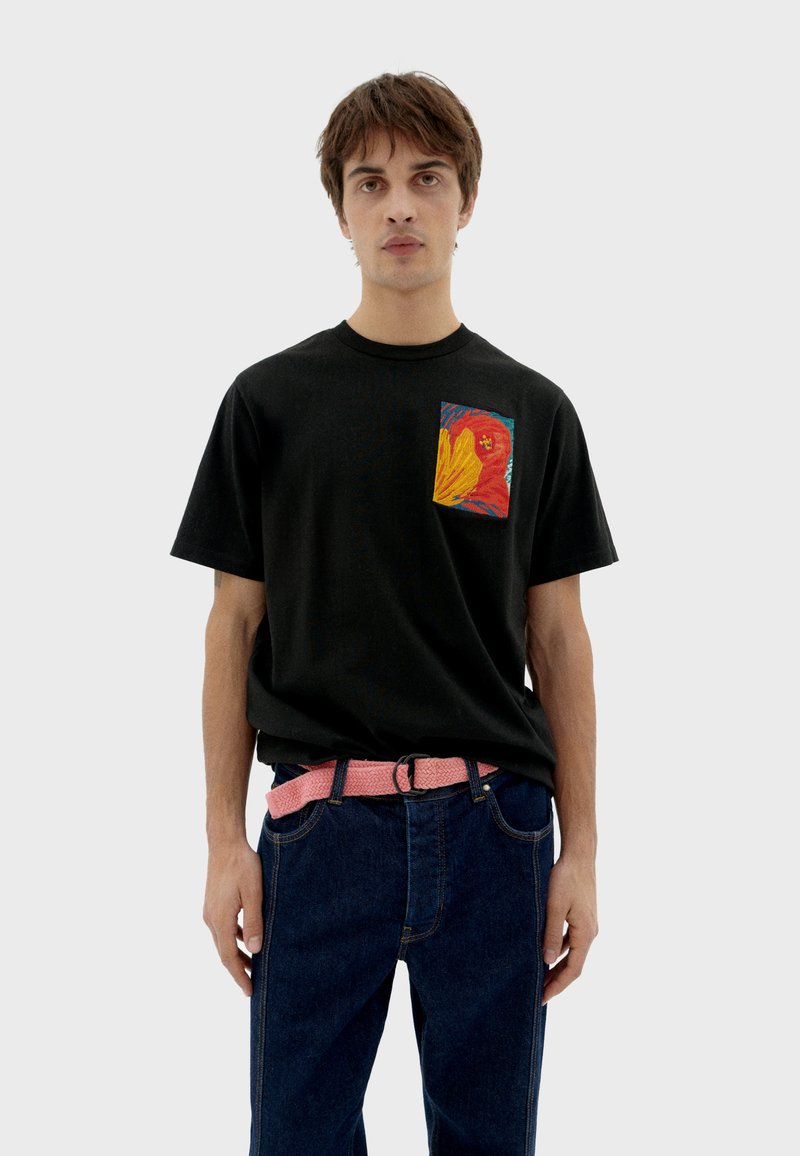 Camiseta negra con un diseño de bolsillo abstracto colorido, combinada con jeans azules y un cinturón de tela rosa con hebilla.