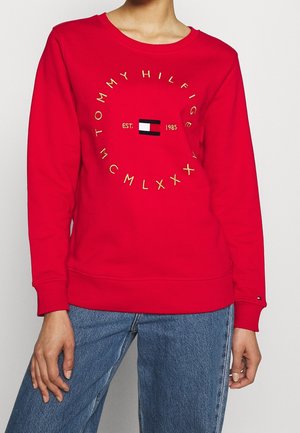Rode sweatshirt met lange mouwen met een cirkelvormige gouden tekst "Tommy Hilfiger", logo in het midden, "EST. 1985," gedragen met blauwe spijkerbroek.