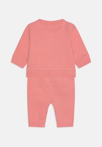 Ružový detský outfit z bavlny, ktorý pozostáva z blúzky s dlhým rukávom, okrúhlym výstrihom a elastickými manžetami, v kombinácii s rovnako elastickými nohavicami.
