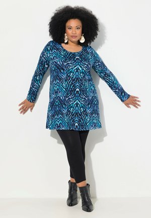 Langsleeve tuniek in een blauw en zwart abstract patroon, gecombineerd met zwarte legging en enkellaarsjes. Het materiaal lijkt zacht met een ontspannen pasvorm.