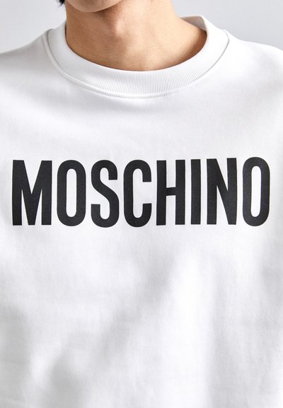 Hvit genser med fet svart "MOSCHINO" tekst på fronten. Stoffet ser mykt ut, med ribbestrikket hals for ekstra detaljer.