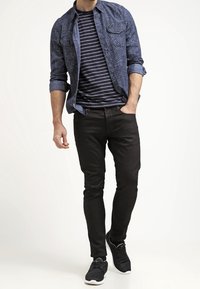 Sininen kuvioitu napitettava paita mustavalkoisen raidoitetun t-paidan päällä, yhdistettynä mustiin slim fit -housuihin ja mustiin tennareihin.