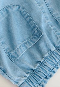 Helles blaues Denim-Gewebe mit strukturiertem Finish, ausgestattet mit zwei aufgesetzten Taschen und einem elastischen Rüschensaum. Sichtbare Nähdetails.