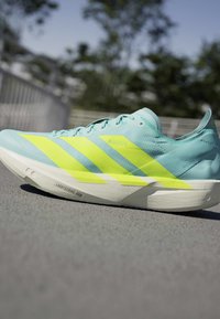 Lyseblå løbesko med tre store gule striber og hvid sål mærket Lightstrike Pro på gråt fortov udendørs.