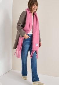 Rosa vævet tørklæde med frynser, båret over en grå frakke og hvid skjorte, kombineret med blå jeans og beige sneakers.