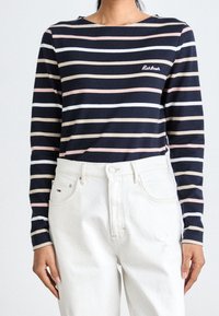 T-shirt à manches longues avec un motif rayé bleu marine et pastel, col rond et logo brodé. Associé à un jean blanc taille haute.