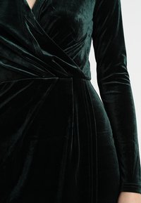 Mørkegrøn fløjlskjole med indpakningsdesign, lange ærmer og intrikate sammensyninger langs siden. Blød tekstur og glans.