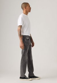 Man met tatoeages in een wit T-shirt, grijze jeans en zwarte sneakers, staande zijwaarts tegen een effen lichte achtergrond.