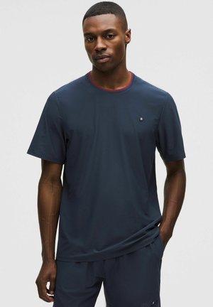 Hombre vistiendo una camiseta deportiva de manga corta azul marino y pantalones cortos a juego, de pie frente a un fondo liso y claro.