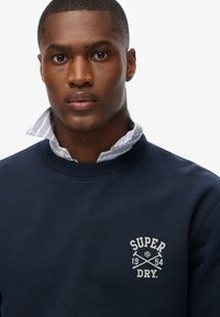 Sudadera navy con cuello acanalado, que presenta un logotipo blanco y un diseño de flechas cruzadas en el pecho. Debajo, se asoma un cuello de camisa a rayas azul y blanco.