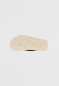 Semelle de chaussure en caoutchouc crème avec surface texturée. Présente un motif diagonal et le nom de la marque « GANT » en lettres majuscules en relief.