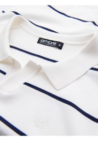 Bílá polo košile s úzkými námořnickými a modrými pruhy, která má žebrovaný límec a vyšitý logo na hrudi. Viditelné štítky.