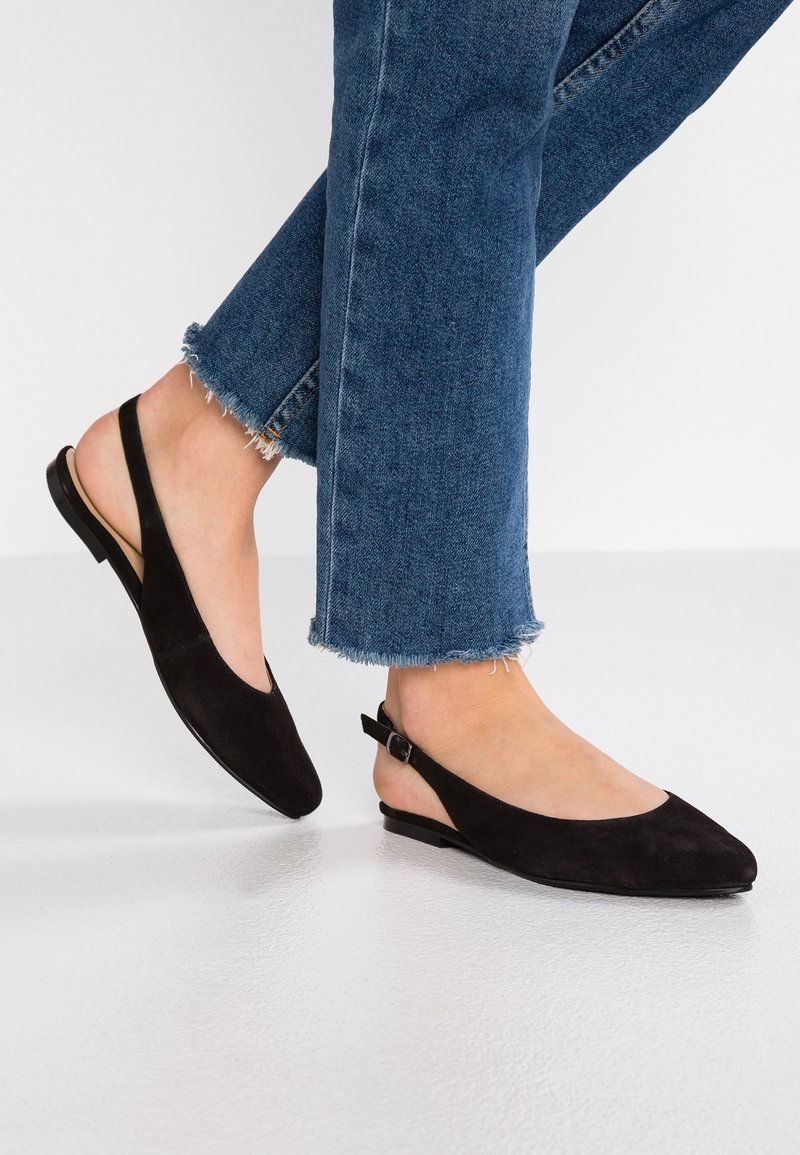Ballerines noires en daim avec un bout pointu, semelle plate et une fine bride de cheville réglable. Portées avec un jean délavé à l'ourlet effiloché.