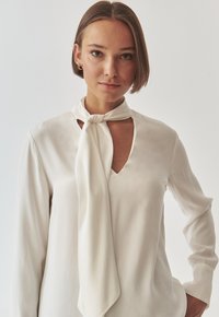 White blouse with a tied scarf detail at the neckline, featuring a subtle sheen and long sleeves with elastic cuffs, smooth texture.