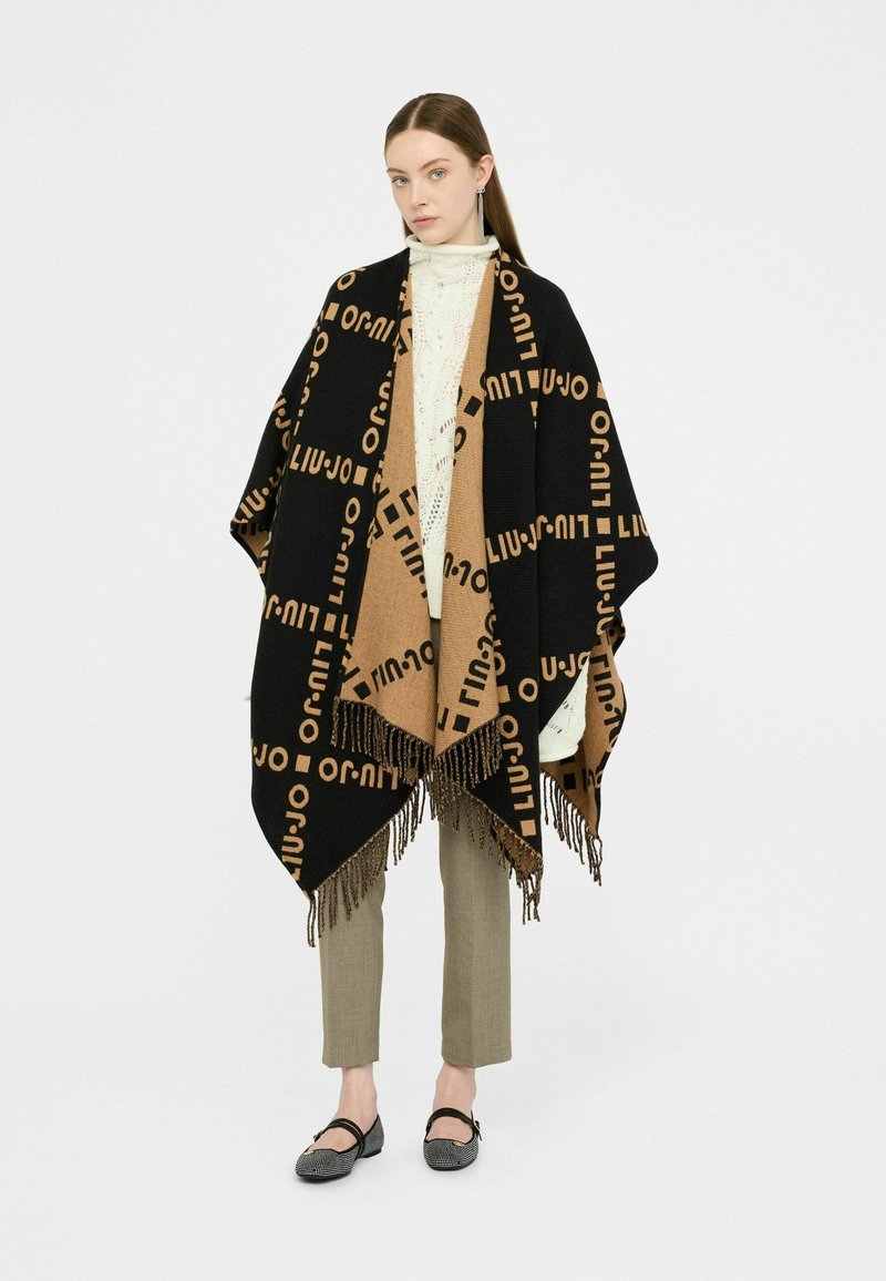 Zwarte en tan poncho met logopatroon, franjes aan de randen, gelaagd over een witte gebreide top en bruine broek, gecombineerd met zwarte platte schoenen.
