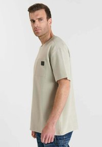 Lichtbeige overhemd met korte mouwen van een gestructureerde stof, voorzien van een voorkeurszak en een vierkante zwarte labeldetail. Draagt bij blauwe jeans.