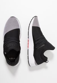 Schwarz-weiße Sport sneakers mit Mesh-Obermaterial, elastischen Riemen und einer dicken weißen Sohle mit roten Akzenten. Gerundete Zehenpartie und strukturierte Details.