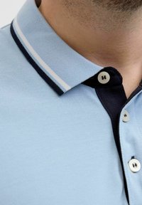 Gaišzils polo krekls ar strukturētu audumu, tumši zila kakla daļa un baltas svītras. Divi pogu neažogos pie kakla ir ar kontrastējošām krāsām.