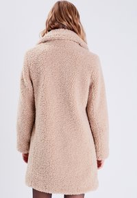 Cappotto finto pelliccia beige con superficie texturizzata e soffice, maniche lunghe e ampio colletto, caratterizzato da una vestibilità ampia e un taglio di lunghezza media.