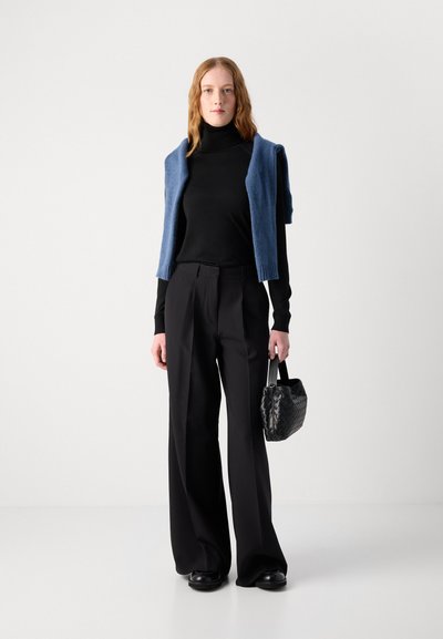 Col roulé noir, pull en mailles bleu drapé sur les épaules, pantalon noir à jambes larges, sac à main noir texturé, chaussures noires, arrière-plan neutre.