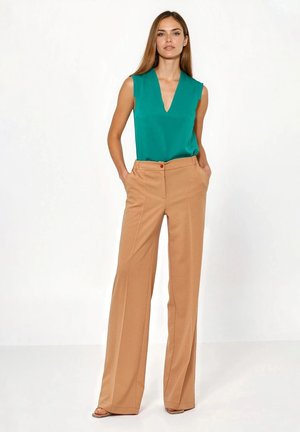 Bluzka bez rękawów w kolorze morskim z dekoltem w serek, zestawiona z wysokim stanem, szerokimi beżowymi spodniami z widocznym czerwonym guzikiem i bocznymi kieszeniami.
