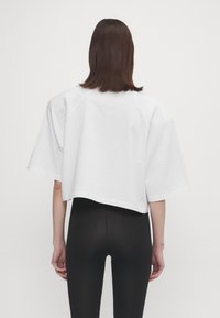 White cropped t-shirt with short, wide sleeves and a loose fit, made of smooth fabric. Worn with black leggings, viewed from the back.