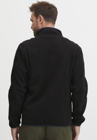 Chaqueta de forro polar negra con cuello alzado, tejido texturizado y ribete azul en la parte inferior. Presenta un detalle de yugo en la parte trasera.