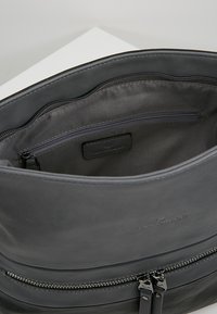 Bolso de cuero gris con una textura suave, cierre con cremallera y bolsillo interior. Presenta una etiqueta con el logo y dos tiradores de cremallera.