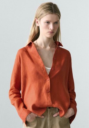 Junge Frau trägt ein lockeres, brennend orangefarbenes Hemd über einem weißen Oberteil, die Hände in den Taschen, vor einem schlichten hellen Hintergrund.