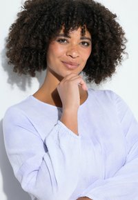 Lichtpaarse blouse met lange mouwen en een geplooide manchetkant. Zachte, gestructureerde stof, gedragen door een persoon die poseert met een bedachtzame uitdrukking.