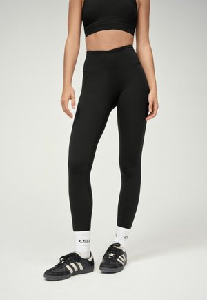 Kvinde iført sorte leggings med høj talje, sort crop top, hvide sokker og sorte Adidas-sneakers står på en ensfarvet baggrund.