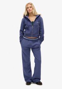 Felpa viola con zip, tasche, lunghezza cropped e cappuccio regolabile, abbinata a pantaloni larghi coordinati con fondo risvoltabile e dettaglio del logo.