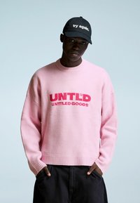 Mlad moški nosi roza pulover z napisom "UNTLD UNTITLED GOODS" in črno kapo z napisom "preizkusi znova." stoji z rokami v žepih.